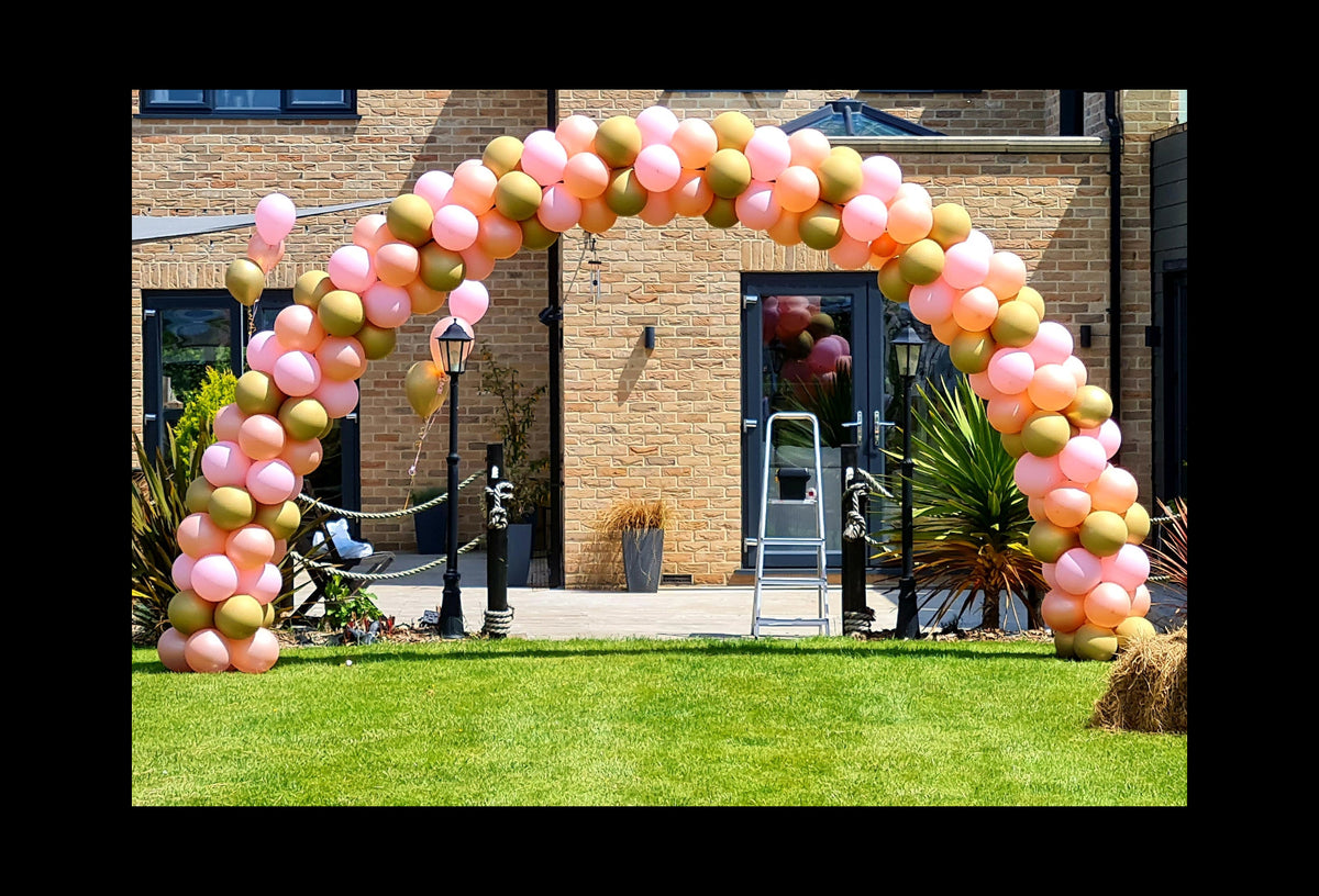 A large balloon arch decoration made of pink, peach, and olive-green balloons, set up on a green lawn in front of a brick building.