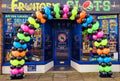 A colorful balloon arch made of black, green, pink, orange, and blue balloons stands in front of a fruit and slot machine shop entrance. The storefront has blue trim, large windows, and a sign reading 'Fruit Shop Slots'.